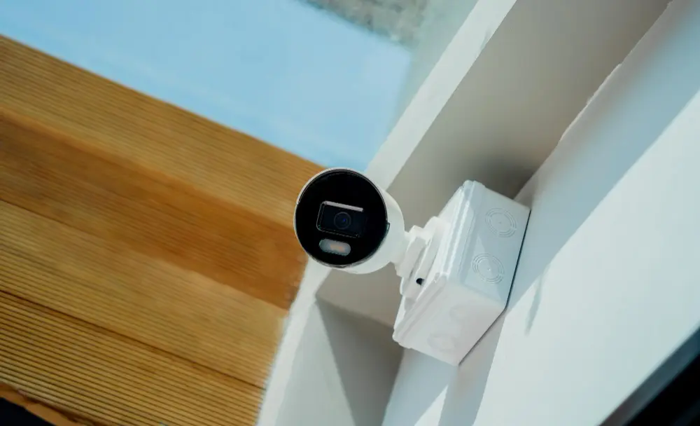 Біла вулична камера відеоспостереження з подвійним об'єктивом і вбудованим прожектором, встановлена під дахом сучасного будинку