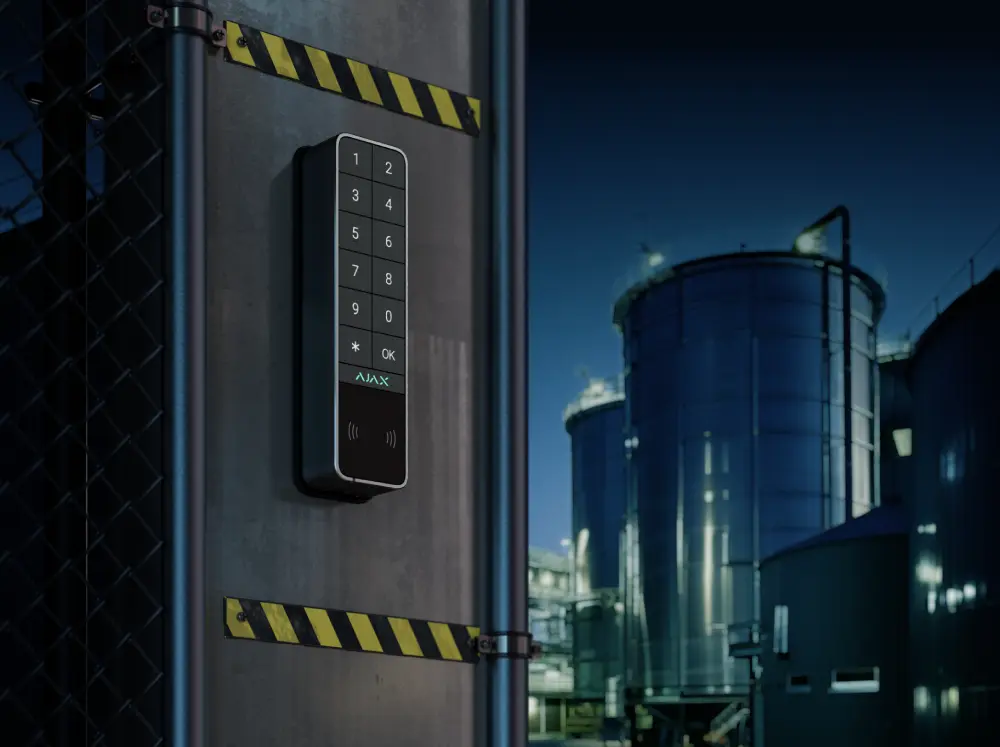 Беспроводная клавиатура Ajax KeyPad Outdoor Jeweller с цифровыми кнопками, RFID-сканером и кнопкой OK, установленная на столбе с ограждением на фоне промышленного объекта с резервуарами ночью