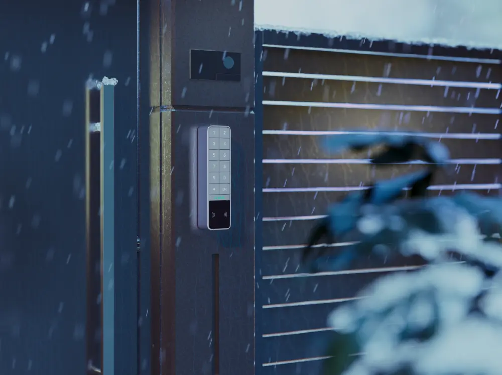 Внешняя беспроводная клавиатура Ajax KeyPad Outdoor Jeweller для управления системой безопасности, с цифровыми кнопками, RFID-сканером и кнопками OK/✱/#, установленная на заборе во время снегопада и дождя