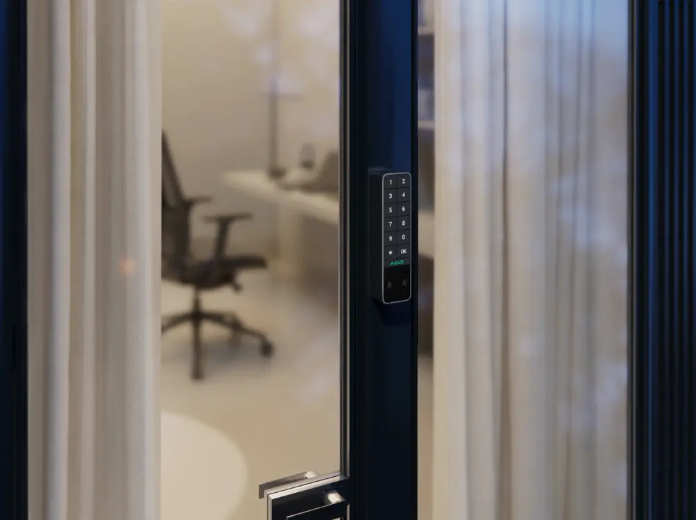 Беспроводная клавиатура Ajax KeyPad Outdoor Jeweller с цифровыми кнопками, RFID-сканером и кнопкой OK, установленная на дверях современного офиса с креслом и шторами на фоне