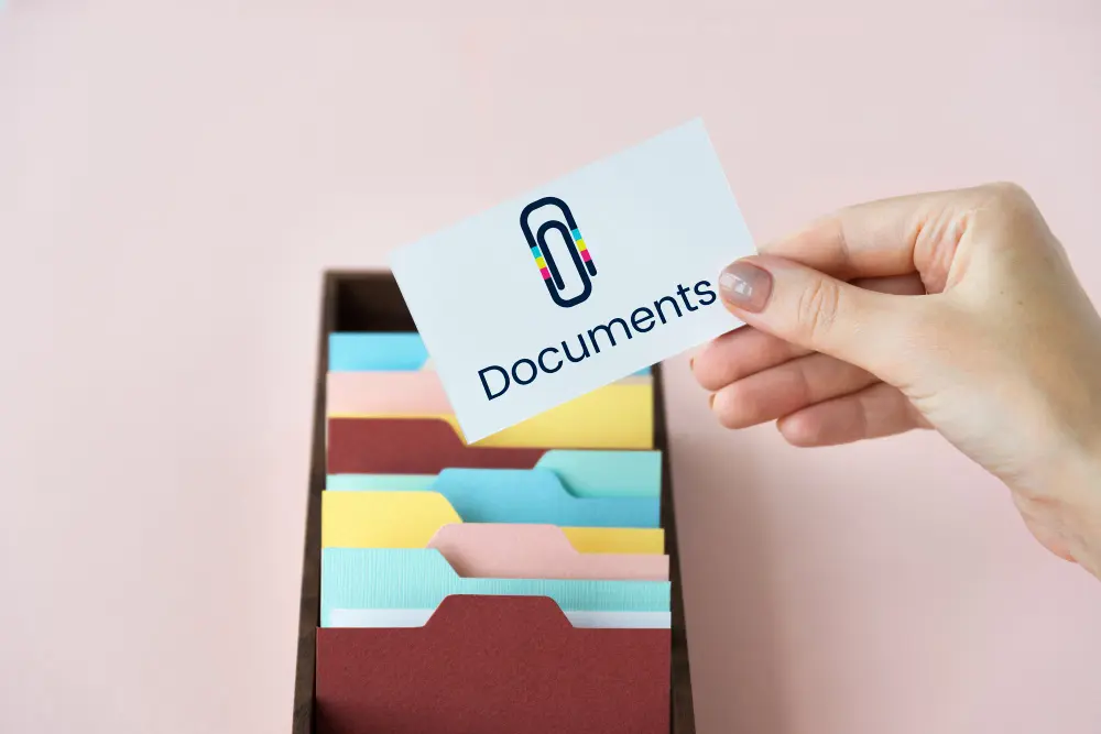 Рука держит карту "Documents" над коробкой с цветными папками на розовом фоне
