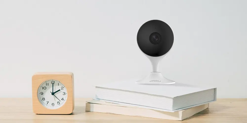 Изображение показывает внутреннюю камеру видеонаблюдения IMOU Cue 2 (IPC-C32EP) на книгах и деревянный будильник со временем 2:54 на столе с минималистичным фоном.